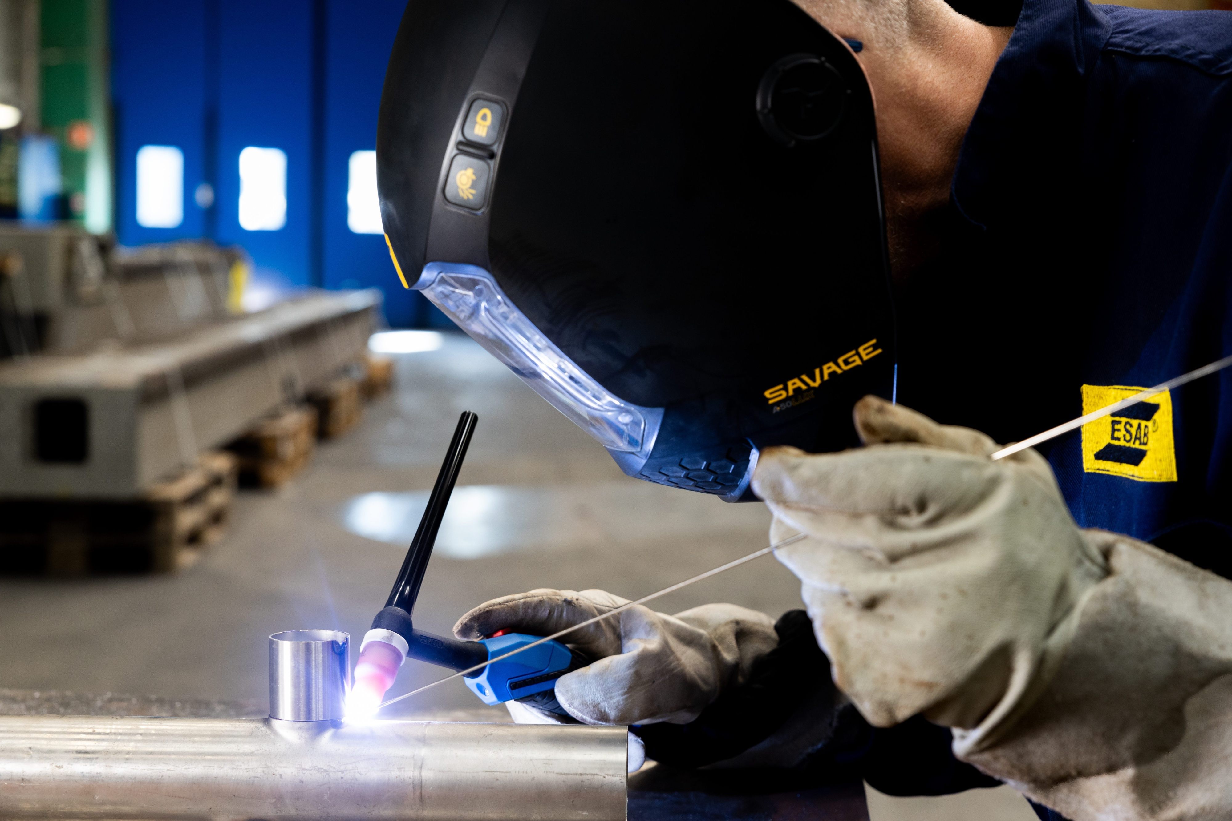 A welder in a helmet works on a metal piece with a TBI tig torch, demonstrating precision and skill.